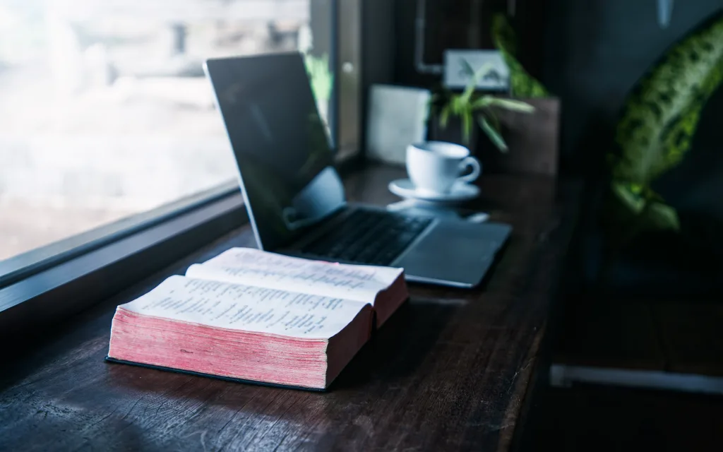 Close up of open Bible on a table next to a laptop and mug of coffee