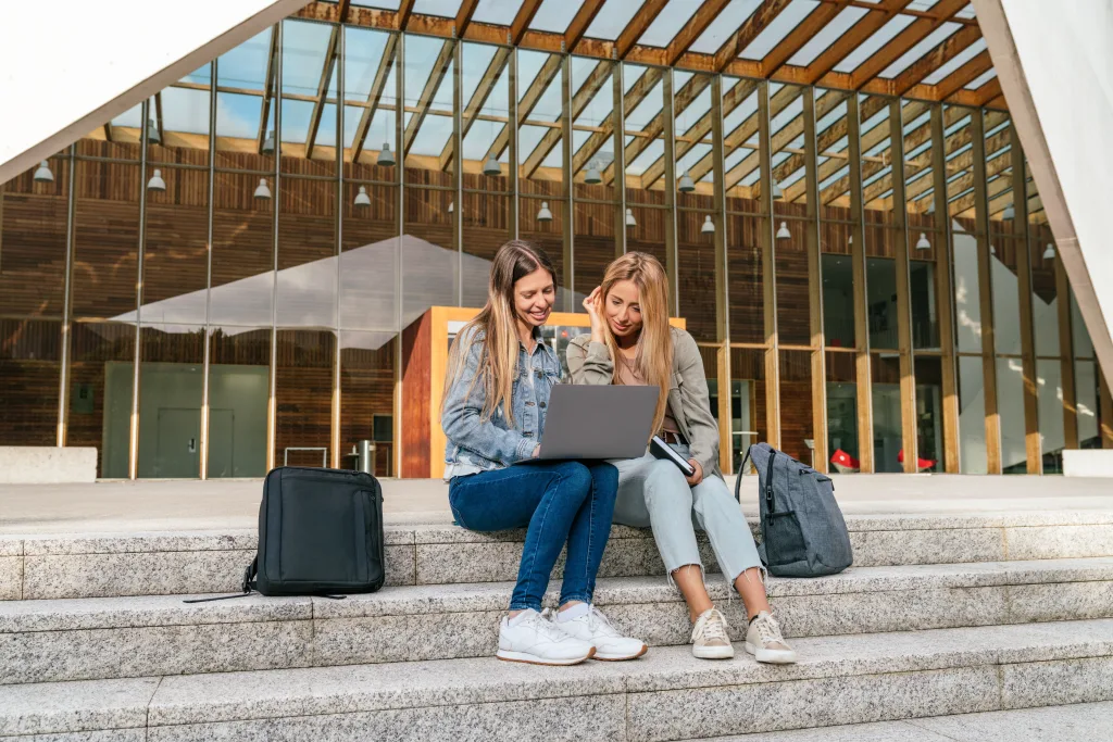 Full length view of two students seated on college campus steps studying on laptop